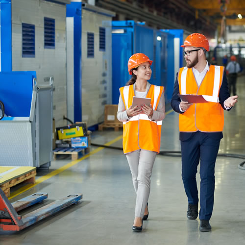 Two workers in safety gear walk through an industrial site, discussing energy-efficient upgrades with tablets in hand.